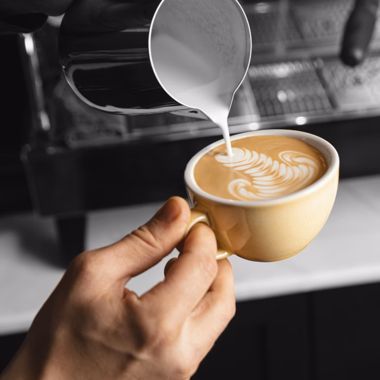 Nahaufnahme von Milch, die in eine Tasse Kaffee mit perfekter Crema gegossen wird.
