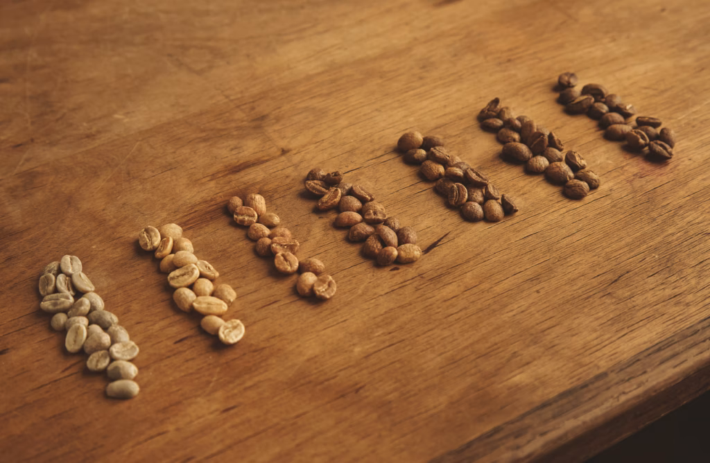 Fünf nebeneinander angeordnete Haufen von Kaffeebohnen in unterschiedlichen Röstgraden – von hellen, fast grünen Bohnen ganz links bis zu intensiv dunklen Röstungen ganz rechts – auf einer hellen Holzoberfläche.