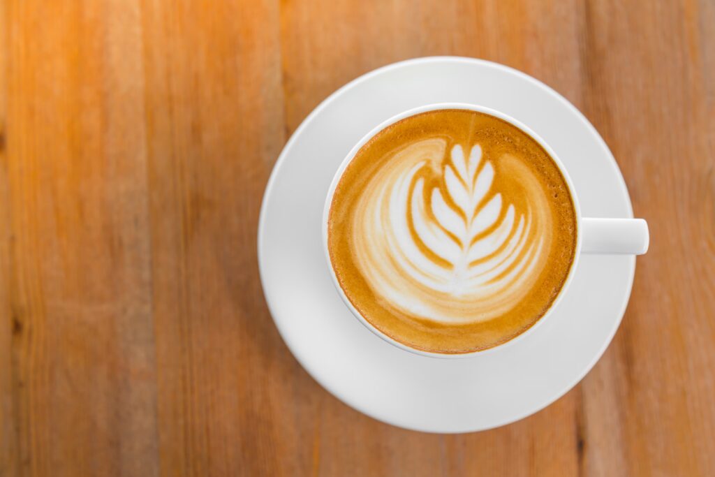Obere Draufsicht auf eine weiße Keramiktasse mit Saucer, die einen Cappuccino mit einer feinen Latte‐Art in Form eines stilisierten Blattmusters zeigt. Die Tasse steht auf einer hellen Holzoberfläche.