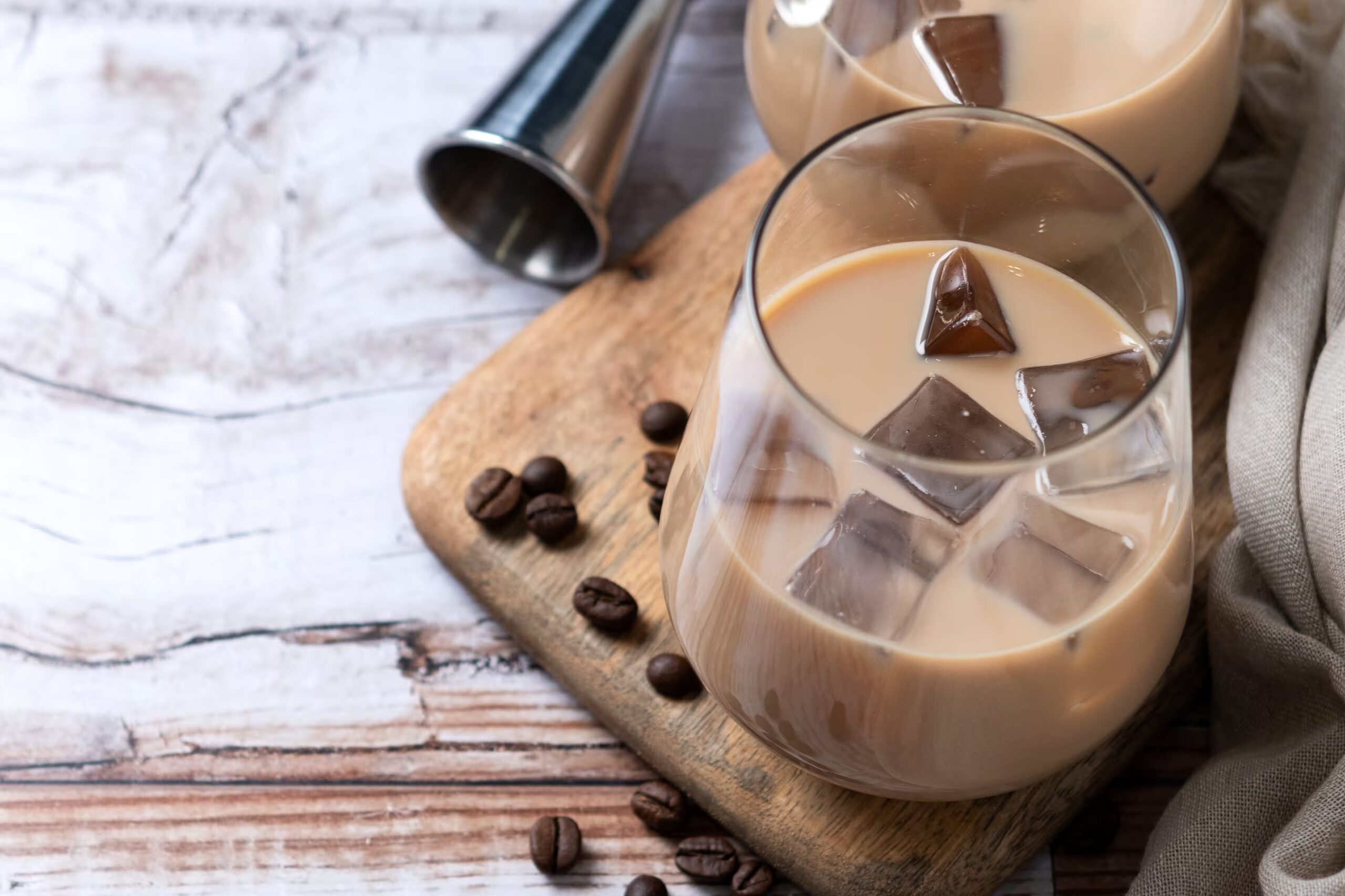Zwei bauchige Gläser mit Eiswürfeln und milchigem Eiskaffee stehen auf einem Holzbrett, daneben liegen verstreute Kaffeebohnen und ein Edelstahl-Jigger; der Hintergrund zeigt eine helle, leicht verwitterte Holzplatte.
