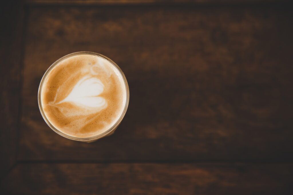 Obere Draufsicht auf ein Glas Latte Macchiato mit Herz‐Latte‐Art. Das Glas steht auf einer dunklen Holztischplatte, der Kontrast zwischen Milchschaum und Kaffee ist gut zu sehen.