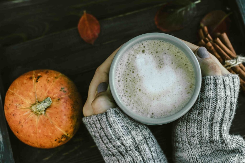 Obere Draufsicht auf eine Person in grauem Strickpullover, die beide Hände um eine graue Keramiktasse Pumpkin Spice Latte hält. Links neben der Tasse liegt ein kleiner, runder Kürbis, rechts sind Zimtstangen und herbstliche Blätter zu sehen.