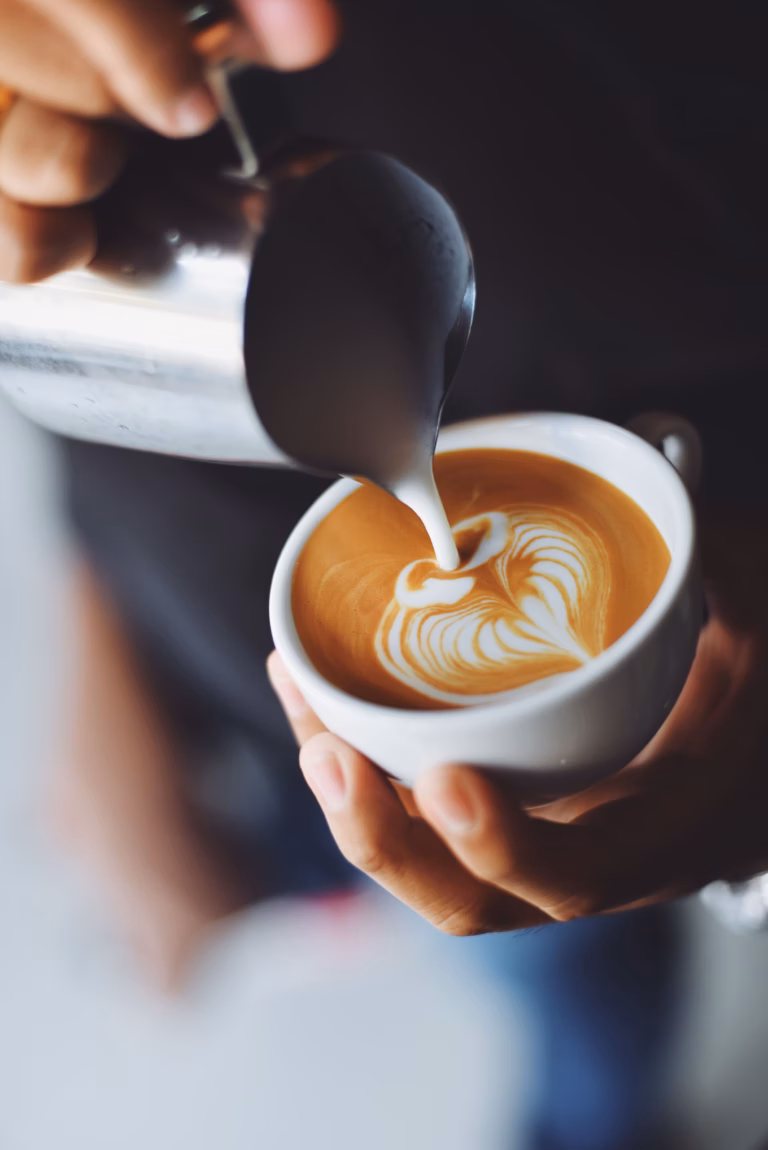 Nahaufnahme von Milch, die in eine Tasse Kaffee mit perfekter latte art gegossen wird.