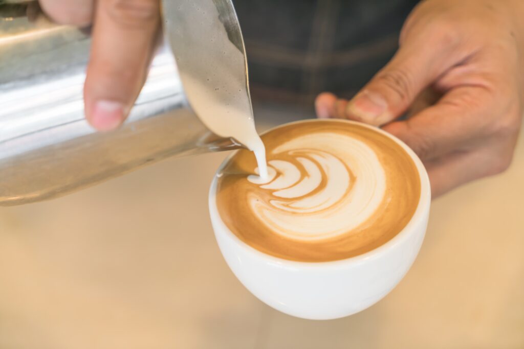 Nahaufnahme einer Person, die mit einer Edelstahl-Milchkännchen feinporigen Milchschaum in eine weiße Kaffeetasse gießt und dabei ein mehrlagiges Latte-Art-Muster in den hellbraunen Kaffee zeichnet.