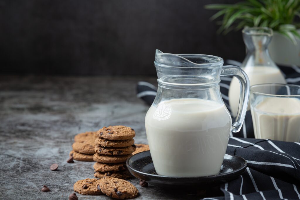 Ein gläserner Milchkrug und ein Glas mit Milch stehen auf einer dunklen, strukturierten Tischplatte. Daneben ist ein Stapel Schokoladenkekse angeordnet, ebenso liegen Kaffeebohnen vereinzelt verstreut.