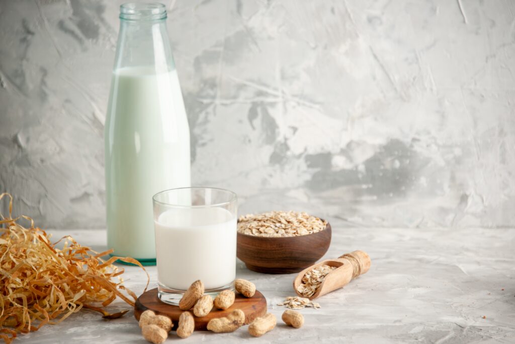 Verschiedene pflanzliche Milchalternativen auf einem hellen Tisch: Eine hohe Glasflasche mit weißer Flüssigkeit, davor ein Glas mit Hafermilch, daneben eine Holzschale mit Haferflocken, eine Schale mit Erdnüssen und verstreute Erdnuss-Schalen.