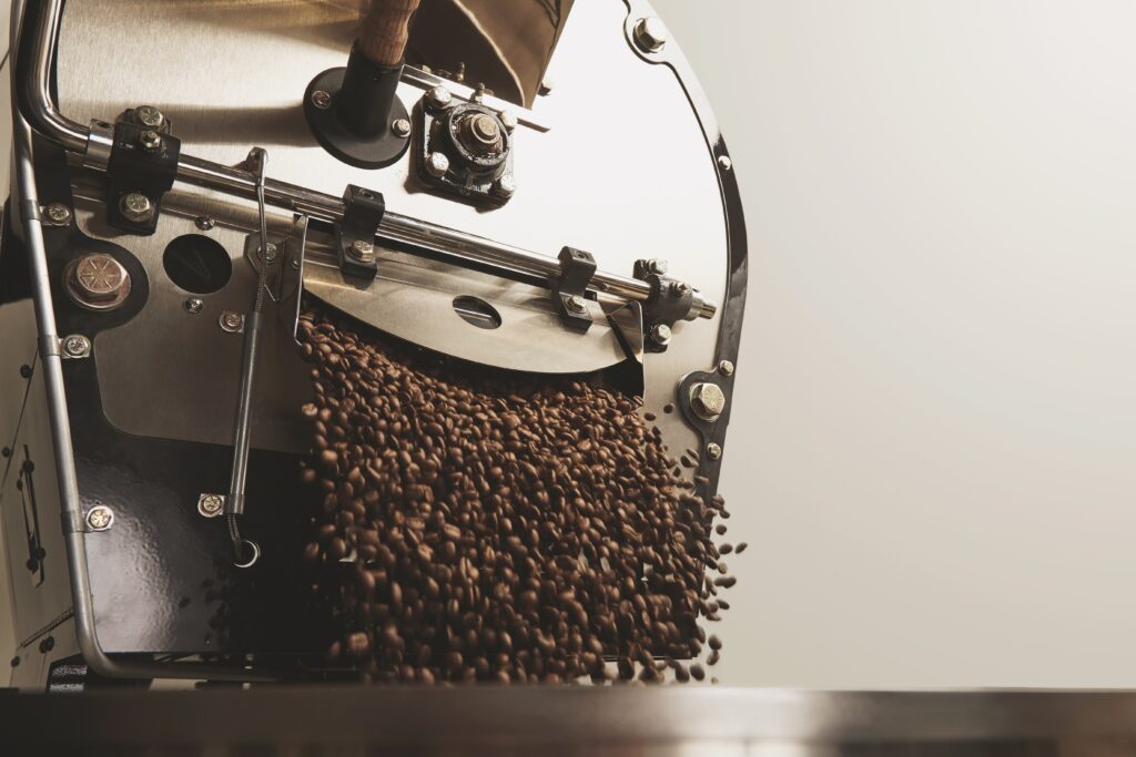 Frisch geröstete Kaffeebohnen fallen aus einem professionellen Trommelröster in eine Edelstahlwanne – Nahaufnahme einer Kaffeerösterei.