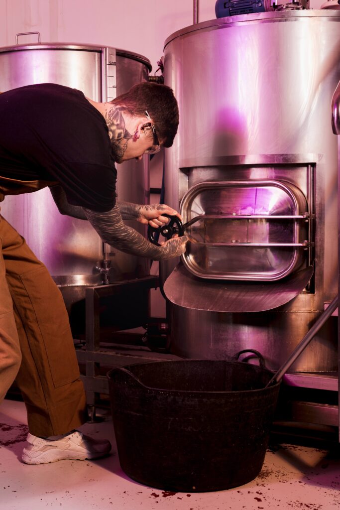 Ein tätowierter Barista öffnet einen großen Röstkessel in einer Kaffeerösterei. Die Szene ist in pink-violettes Licht getaucht, die Umgebung wirkt industriell und modern.